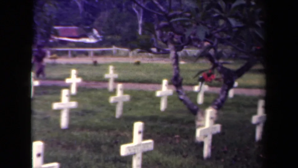 1940s Papua New Guinea 8mm frame of white cross grave markers partly framed by a dark tree, with a path and foliage beyond.