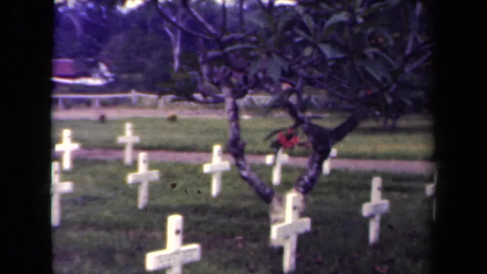 1940s Papua New Guinea 8mm frame showing rows of white wooden crosses, green lawn, and a dark-branched tree with red flowers.