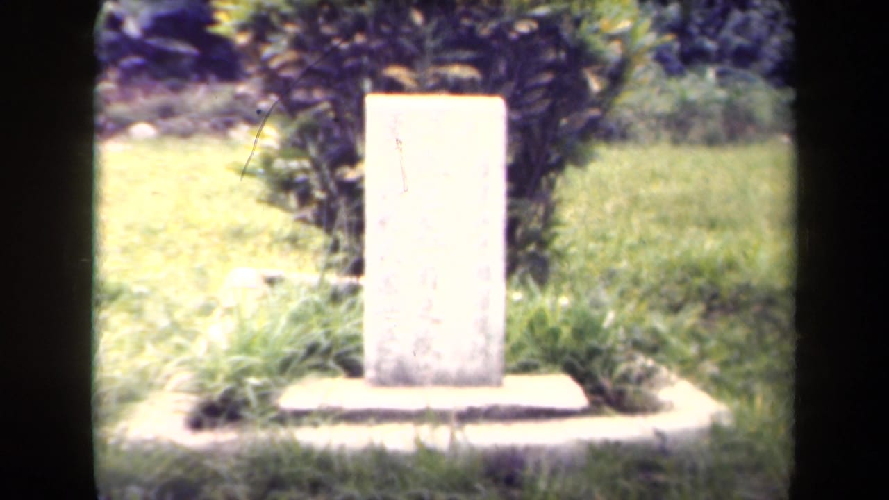 1940s Papua New Guinea footage appears to show a pale memorial stone set in grass with shrubs behind it.