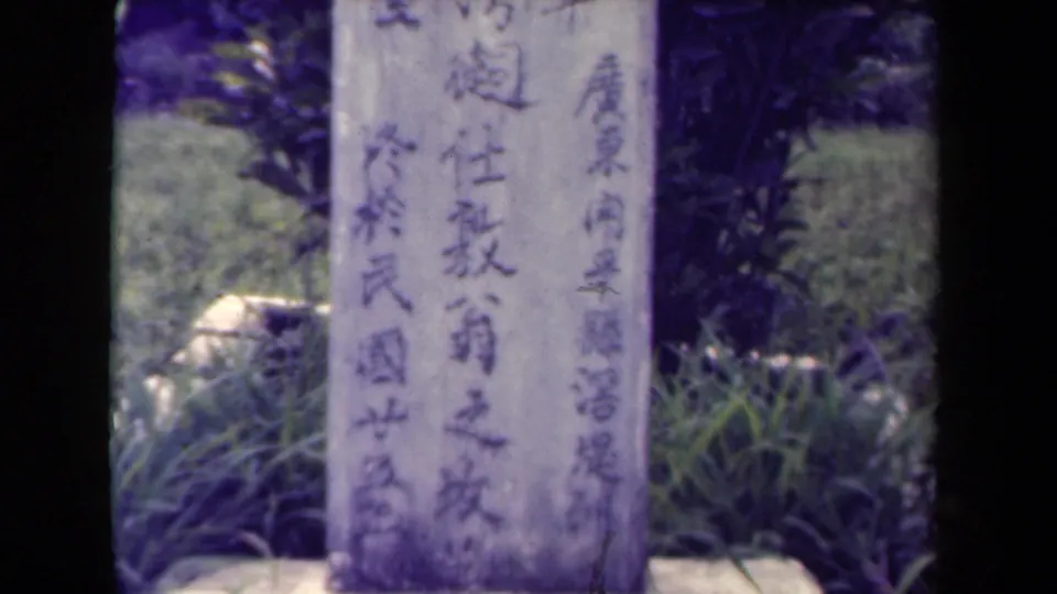 1944 Papua New Guinea 8mm frame showing a weathered stone marker with Chinese characters set amid grass and leafy vegetation.