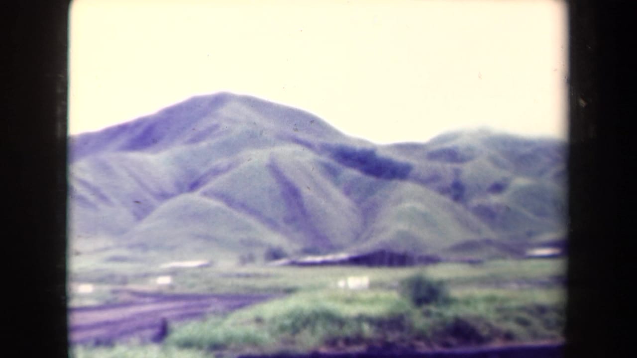 1944 8mm footage from Papua New Guinea showing green mountain slopes, a pale sky, grassland, and an unpaved road in the foreground.