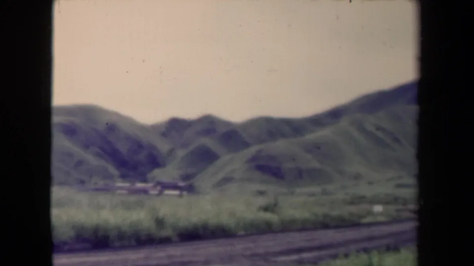 Soft 1944 8mm view in Papua New Guinea, with grassy foreground, low hills, pale sky, and a rough road along the bottom edge.