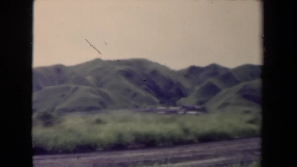 1944 Papua New Guinea landscape frame showing green ridges, grassland, small distant structures, and a dirt road in soft 8mm color.