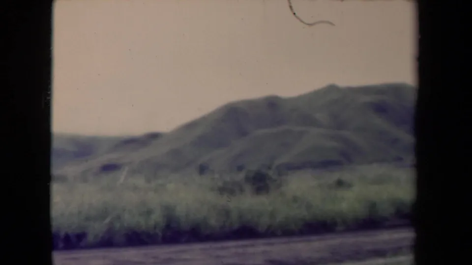 Archival 8mm view of Papua New Guinea hills with grass in the foreground and a road at the bottom of frame.