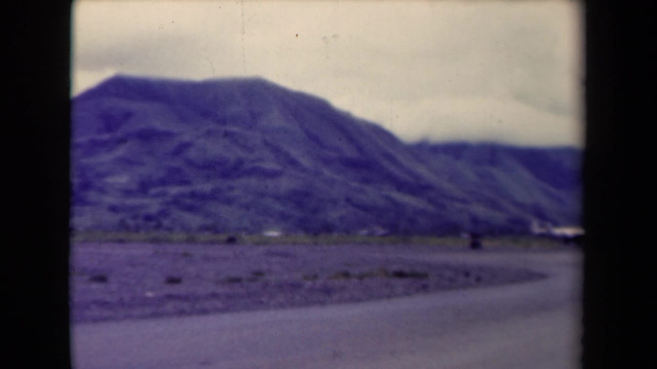 1944 Papua New Guinea 8mm frame showing a curving paved road, scrubby roadside, distant mountains, and overcast sky.