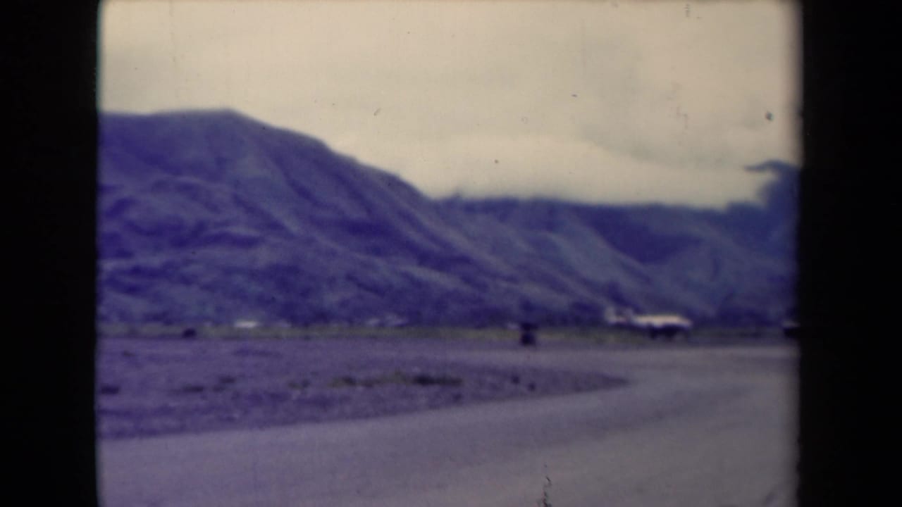 1944 Papua New Guinea roadside view with rugged mountains, a curving road, muted color, and small distant built forms.