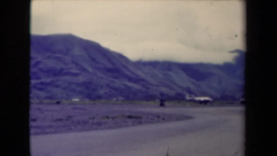 1944 Papua New Guinea roadside view with rugged mountains, a curving road, muted color, and small distant built forms.