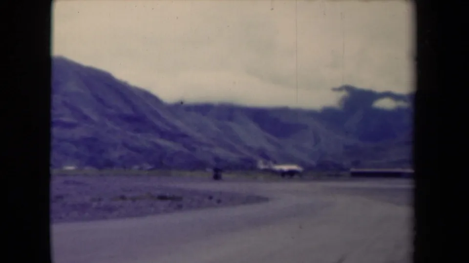 1944 Papua New Guinea 8mm frame showing a road crossing the foreground toward mountains and small distant buildings.