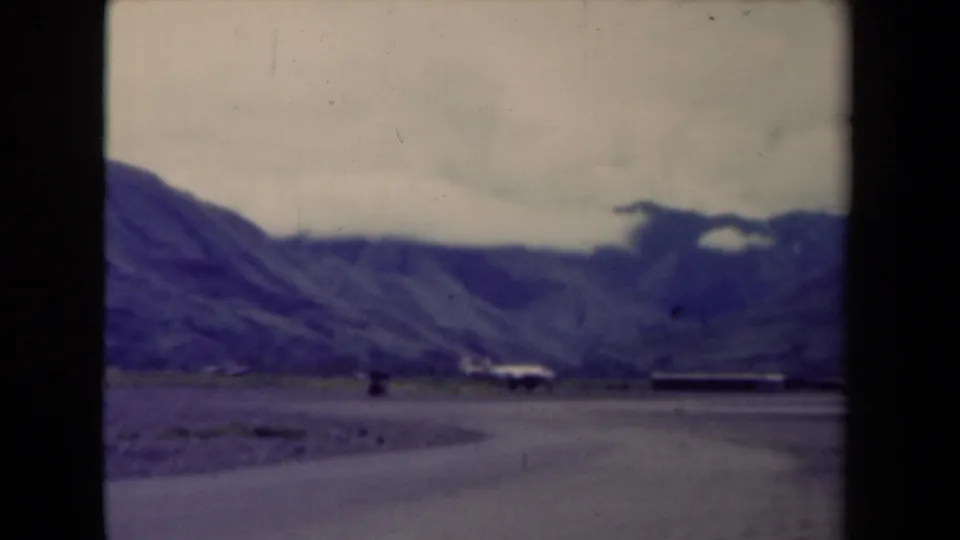 1944 Papua New Guinea landscape frame with a curving road, mountain backdrop, cloudy sky, and small settlement-like forms.