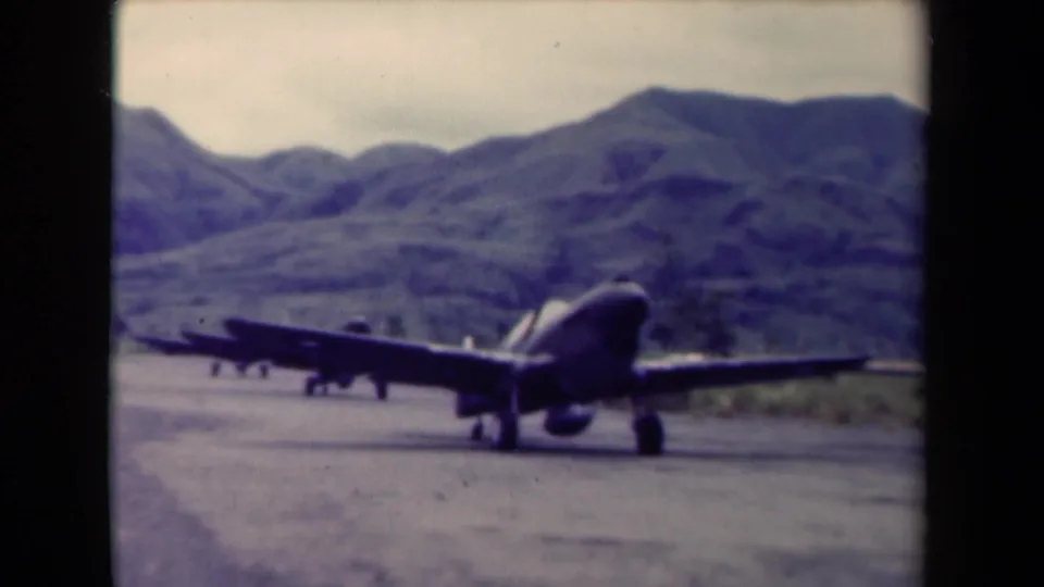 A propeller military aircraft appears on a runway in mountainous Papua New Guinea, with another aircraft faintly visible nearby.