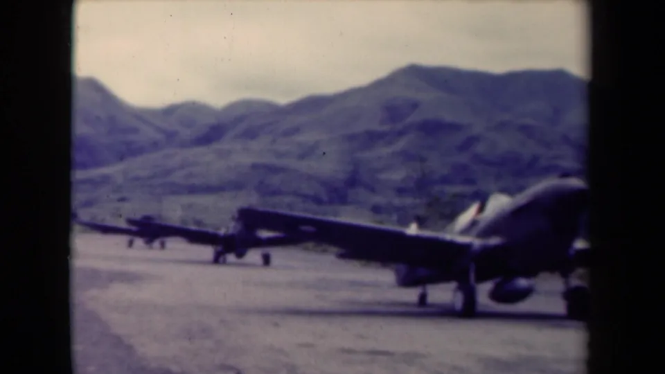 The 8mm frame shows a military aircraft passing close to camera on a Papua New Guinea mountain runway, with another plane farther back.