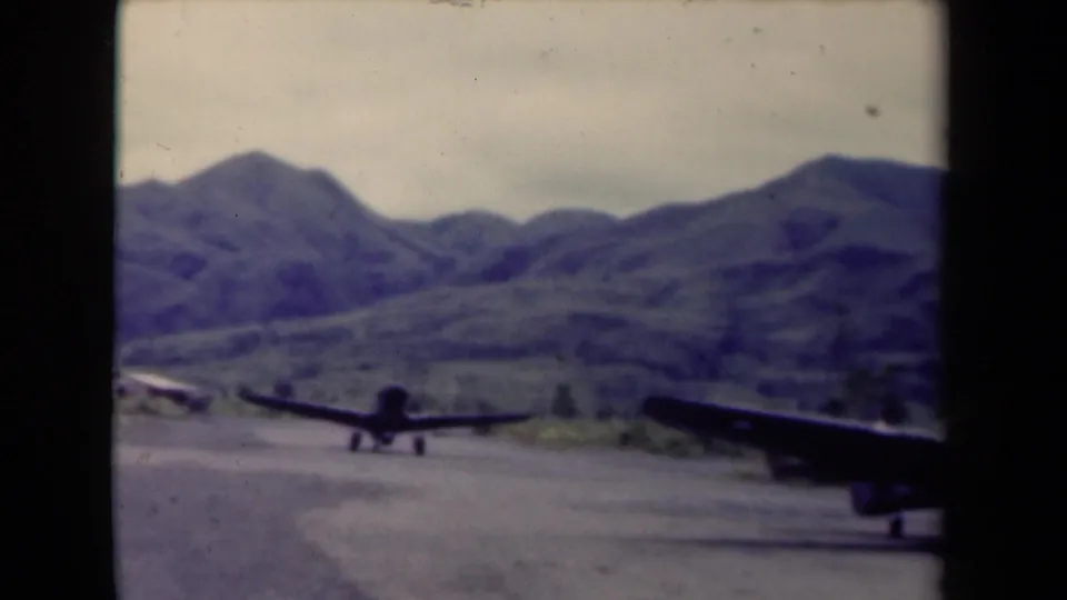 1944 Papua New Guinea 8mm frame showing two aircraft on a rugged airstrip backed by mountainous jungle terrain.
