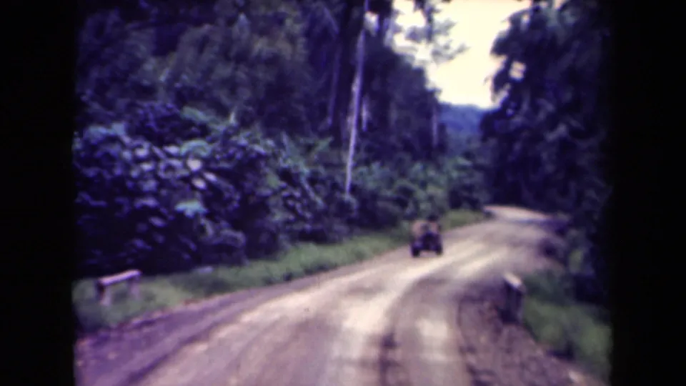In the 1944 Papua New Guinea clip, the truck continues away on a pale unpaved road through thick tropical vegetation.