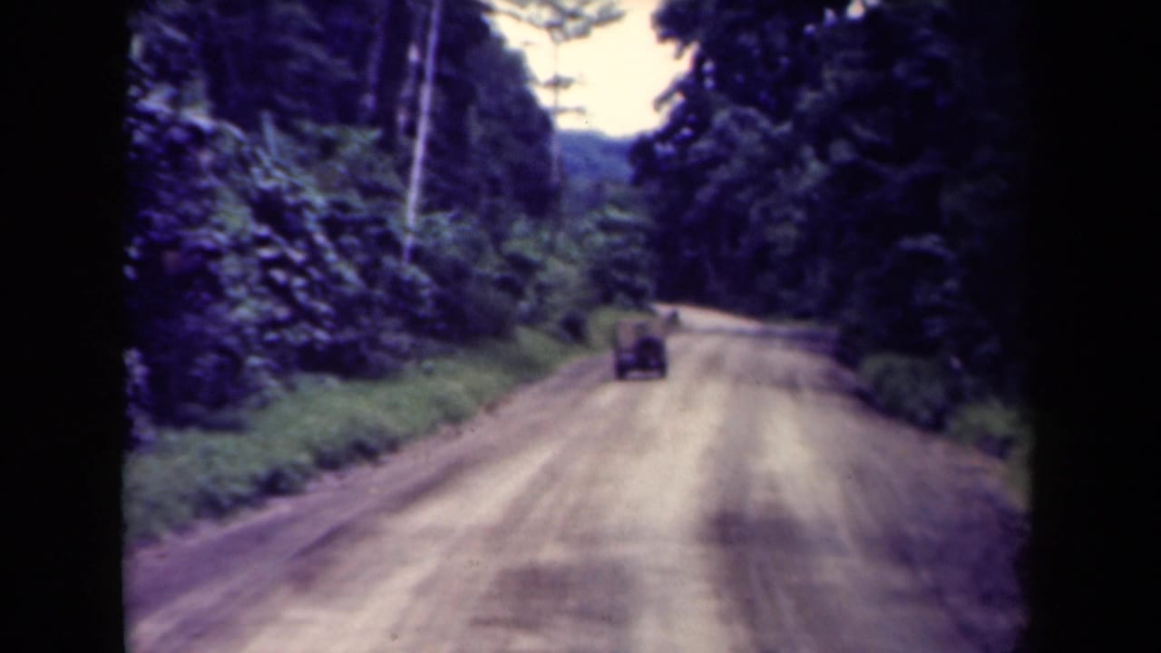 1940s Papua New Guinea 8mm frame with a truck moving away along a pale dirt road enclosed by tropical forest.