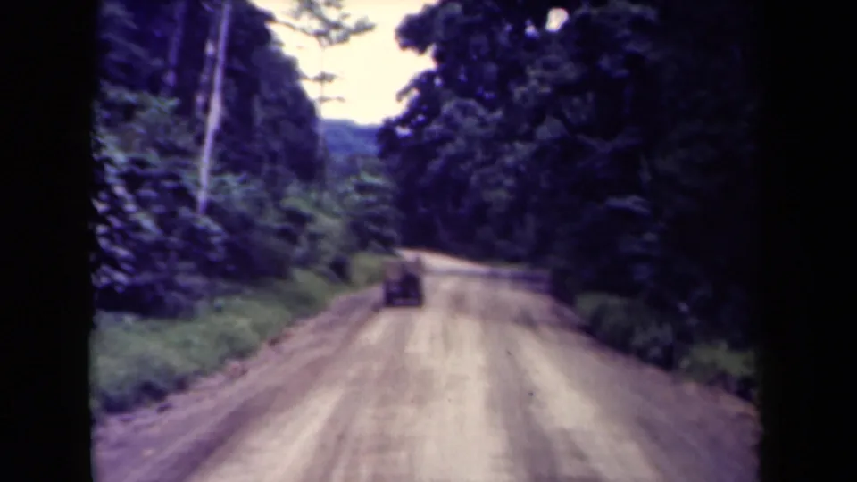 1940s Papua New Guinea 8mm frame showing a small truck centered on a muddy-looking road through dense tropical vegetation.