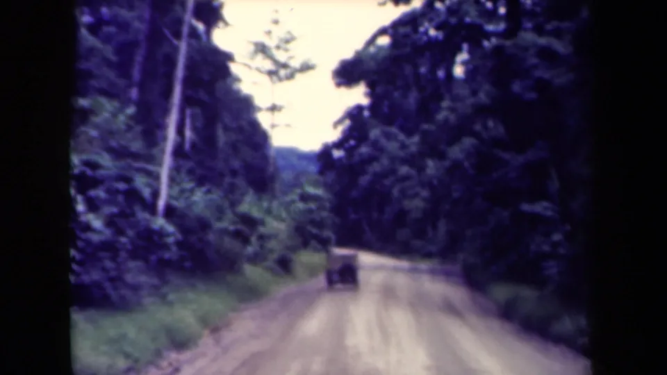 1940s Papua New Guinea 8mm frame of a truck driving away on an unpaved road lined with dense jungle and tall trees.