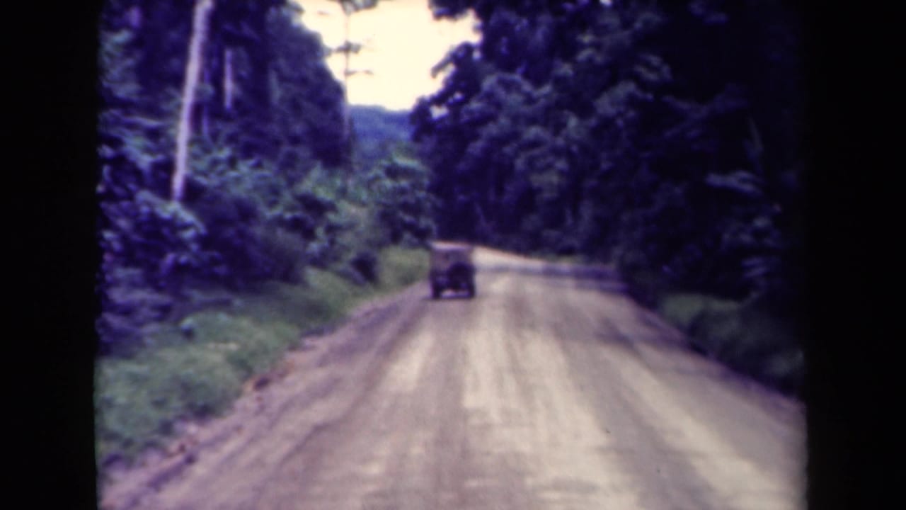 1944 8mm home-movie frame from Papua New Guinea showing a classic car receding along an unpaved forest road.