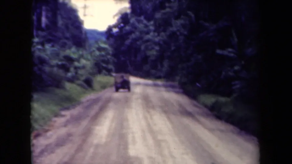 Vintage 8mm frame of a car moving away on a rural Papua New Guinea road with thick forest on both sides.