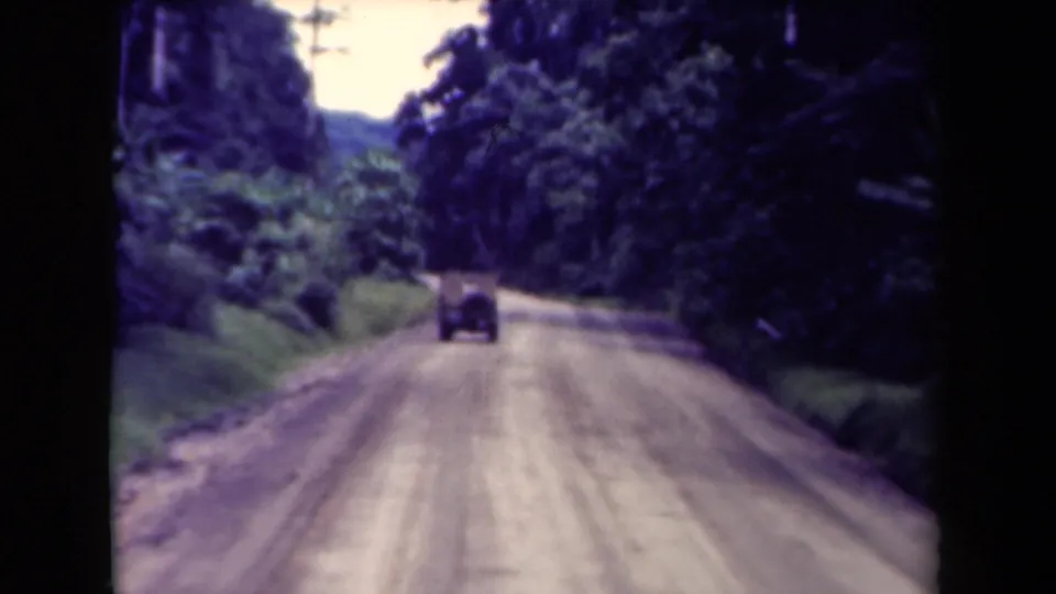 1940s Papua New Guinea travel footage frame showing a small car ahead on a dusty rural road through tropical growth.