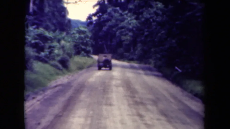 8mm home-movie still from Papua New Guinea with a classic car approaching a bend on a tropical forest dirt road.
