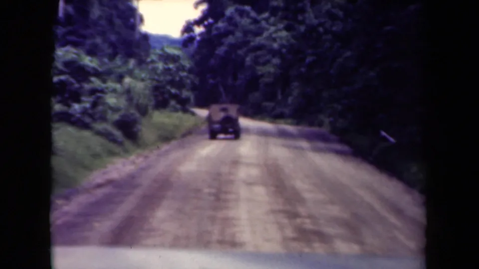 1944 Papua New Guinea 8mm frame showing a jeep ahead on an unpaved road through thick tropical foliage.