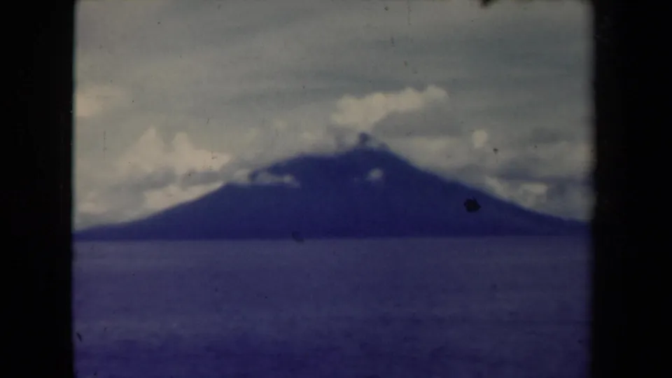 1944 Papua New Guinea 8mm view of a distant mountain or island across the sea beneath a cloudy sky, with blue tint and film wear.