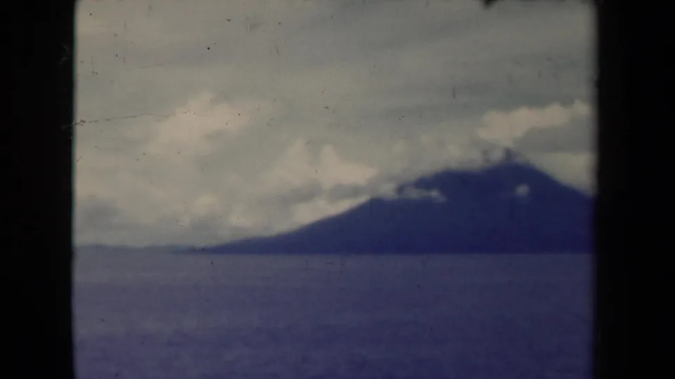 1944 Papua New Guinea coastal landscape frame showing a dark mountain beyond open water, cloudy sky, and visible archival film grain.