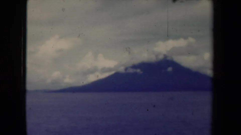 Soft 8mm 1944 Papua New Guinea landscape with a distant mountain across the sea, low clouds, film grain, and dust marks.