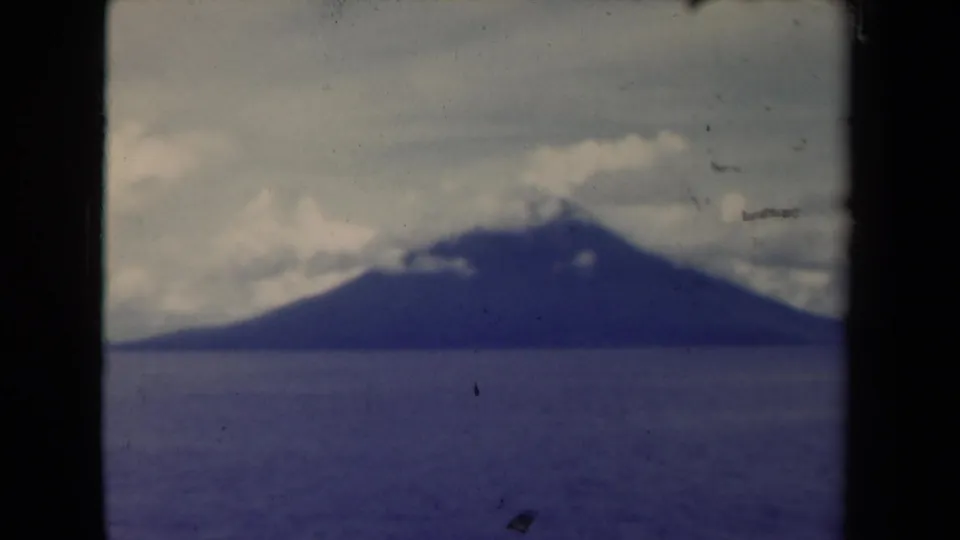 Final 1944 Papua New Guinea 8mm frame of a distant mountain over blue water, with cloud cover, grain, dust, and dark film edge visible.