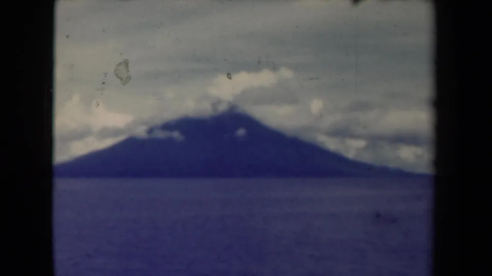 A steady 1940s Papua New Guinea view of a mountain beyond water, with soft focus and visible film wear.