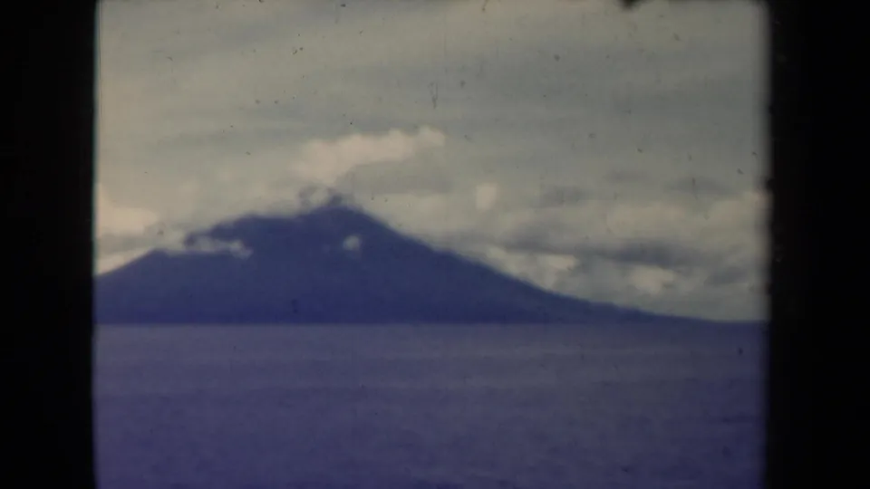 Papua New Guinea home-movie frame from 1944 showing a cloud-wrapped mountain across still water with blue-purple film cast.