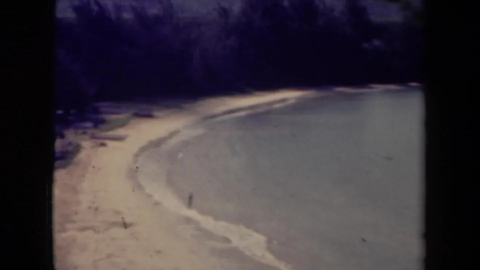 1944 Kauai home-movie frame showing a pale beach, gentle surf, wooded shoreline, and distant mountains across the water.