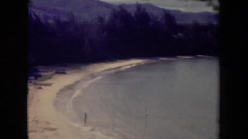 1944 Kauai coastal view with sandy shore, calm bay, distant mountains, and a small person standing near the surf.