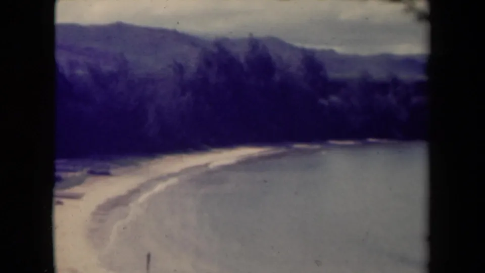 Vintage 8mm Kauai frame with a pale beach curving along the sea below dark trees and hazy mountains.