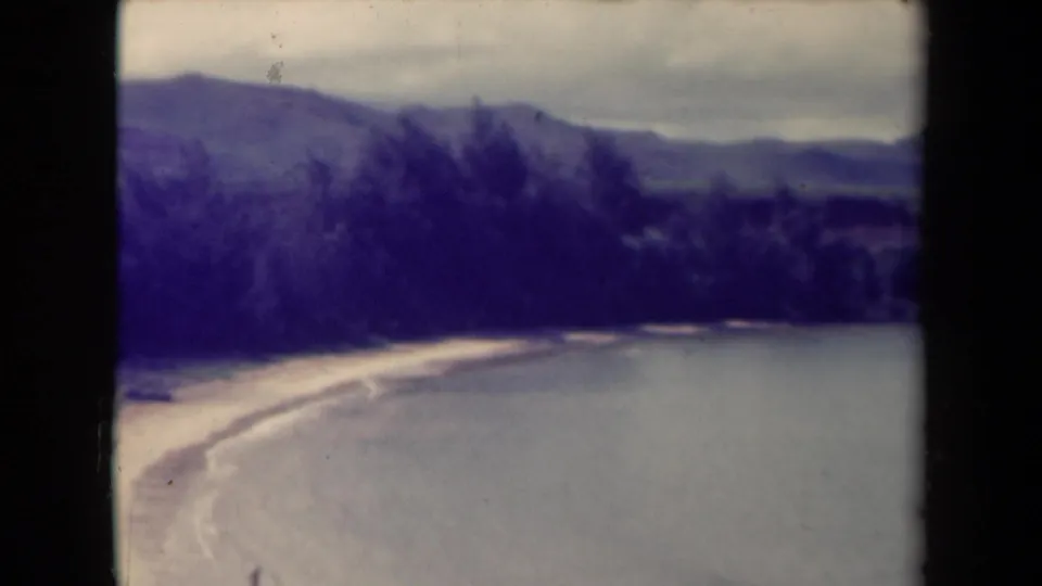 1944 Kauai coastal landscape with mountains, wooded shoreline, pale sand, and smooth water in a faded 8mm frame.
