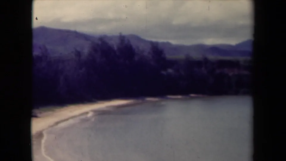 1944 Kauai 8mm frame showing a calm water view bordered by pale shore, dark vegetation, distant mountain ridges, and overcast sky.