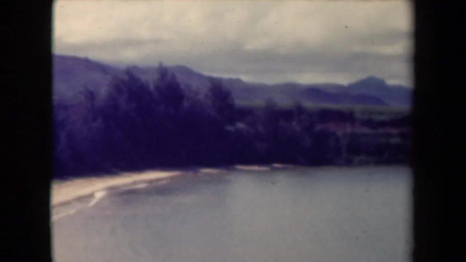1944 Kauai home-movie frame with broad water, a light shoreline edge, dark trees, and distant mountains softened by film grain.