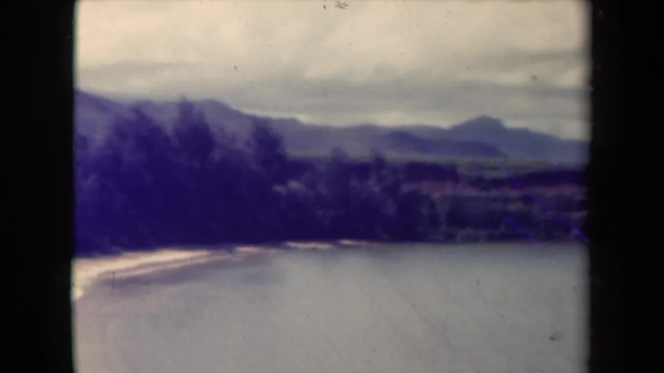 1944 Kauai landscape frame showing still water in the foreground, a sandy shore, dark tropical vegetation, and hazy mountain forms beyond.