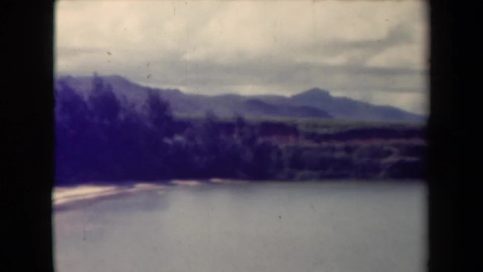 1944 Kauai 8mm frame with calm water below a pale shore, dark trees, distant mountain ridges, and visible home-movie frame borders.