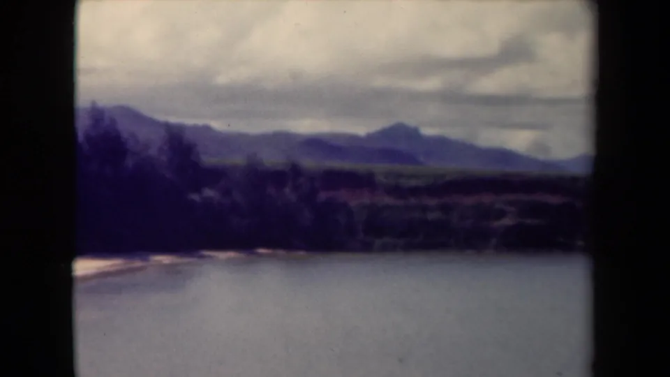 1944 Kauai home-movie frame showing still water, distant wooded hills, and a cloudy sky in soft 8mm color.