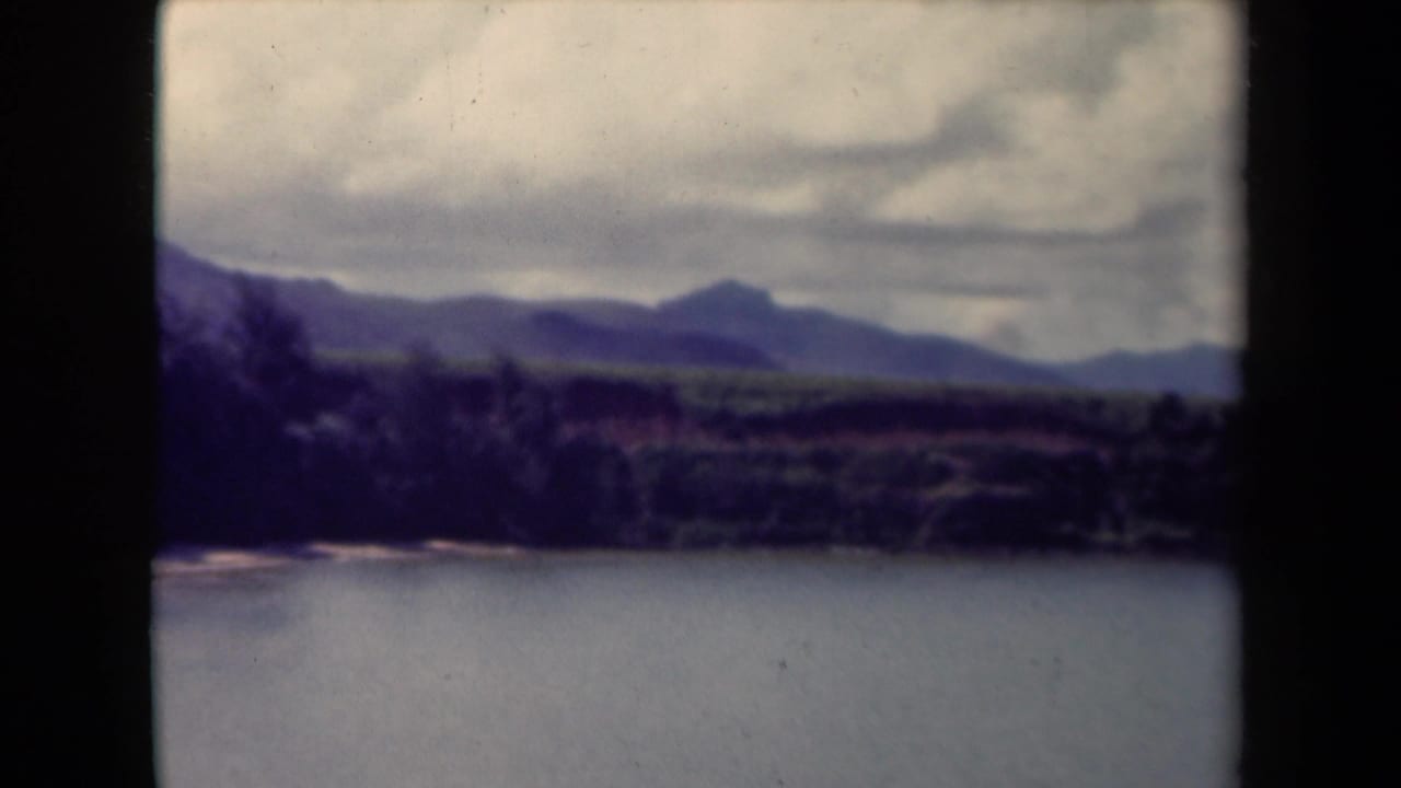 1944 Kauai landscape frame with water in the foreground, a wooded shoreline, distant hills, and cloud cover.