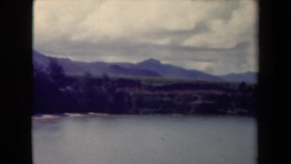 1944 Kauai landscape frame with water in the foreground, a wooded shoreline, distant hills, and cloud cover.
