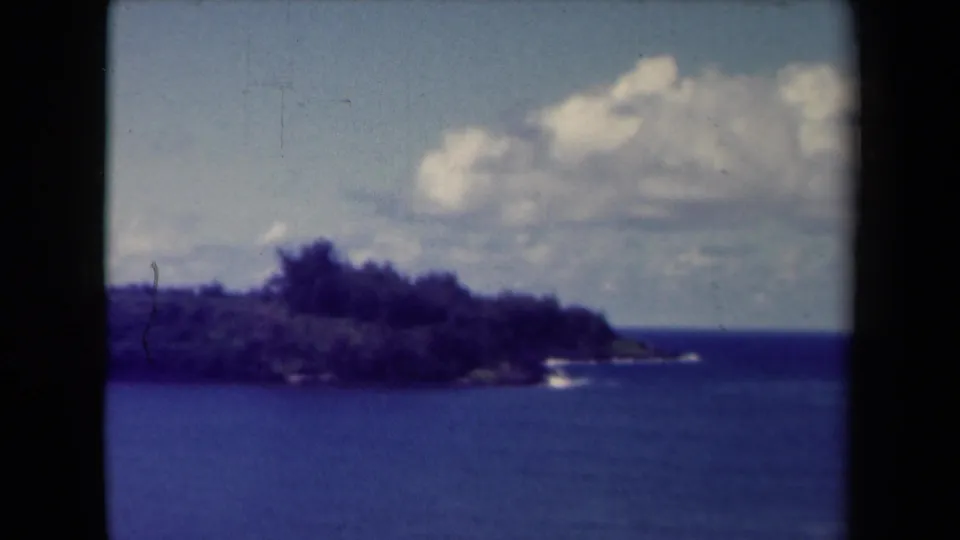 1944 Kauai home-movie frame showing a dark wooded coastal point, blue ocean, and bright clouds overhead.