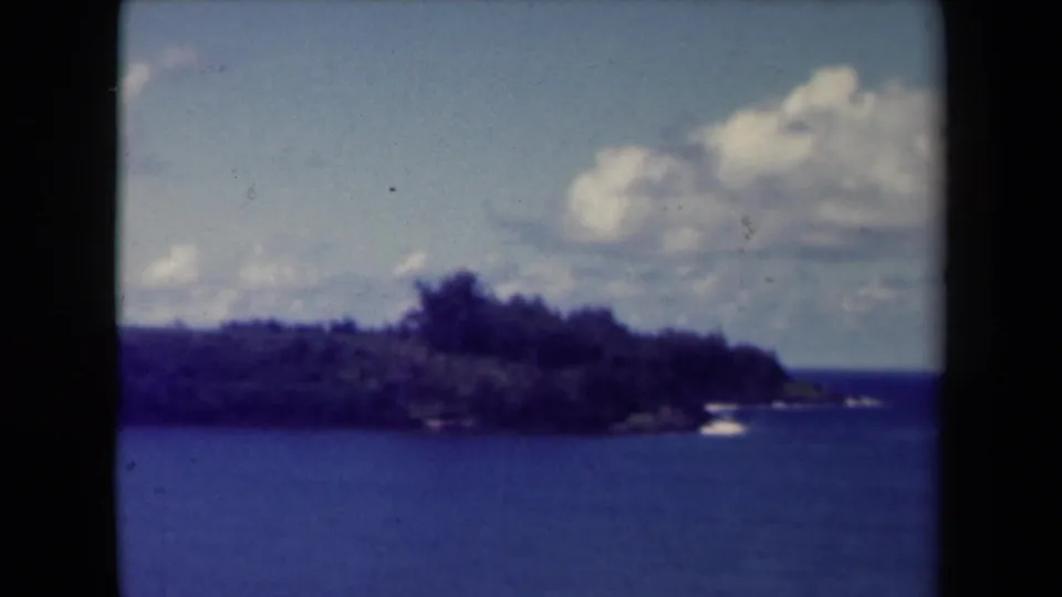 1944 Kauai frame showing open water, a tree-covered coastal edge, and sunlit clouds in soft 8mm texture.