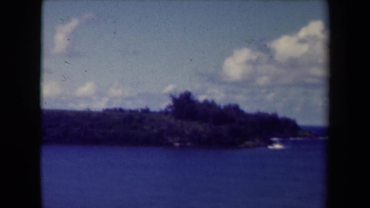 1944 Kauai 8mm frame showing calm blue water, a dark green shoreline, distant low hills, and a partly cloudy sky.