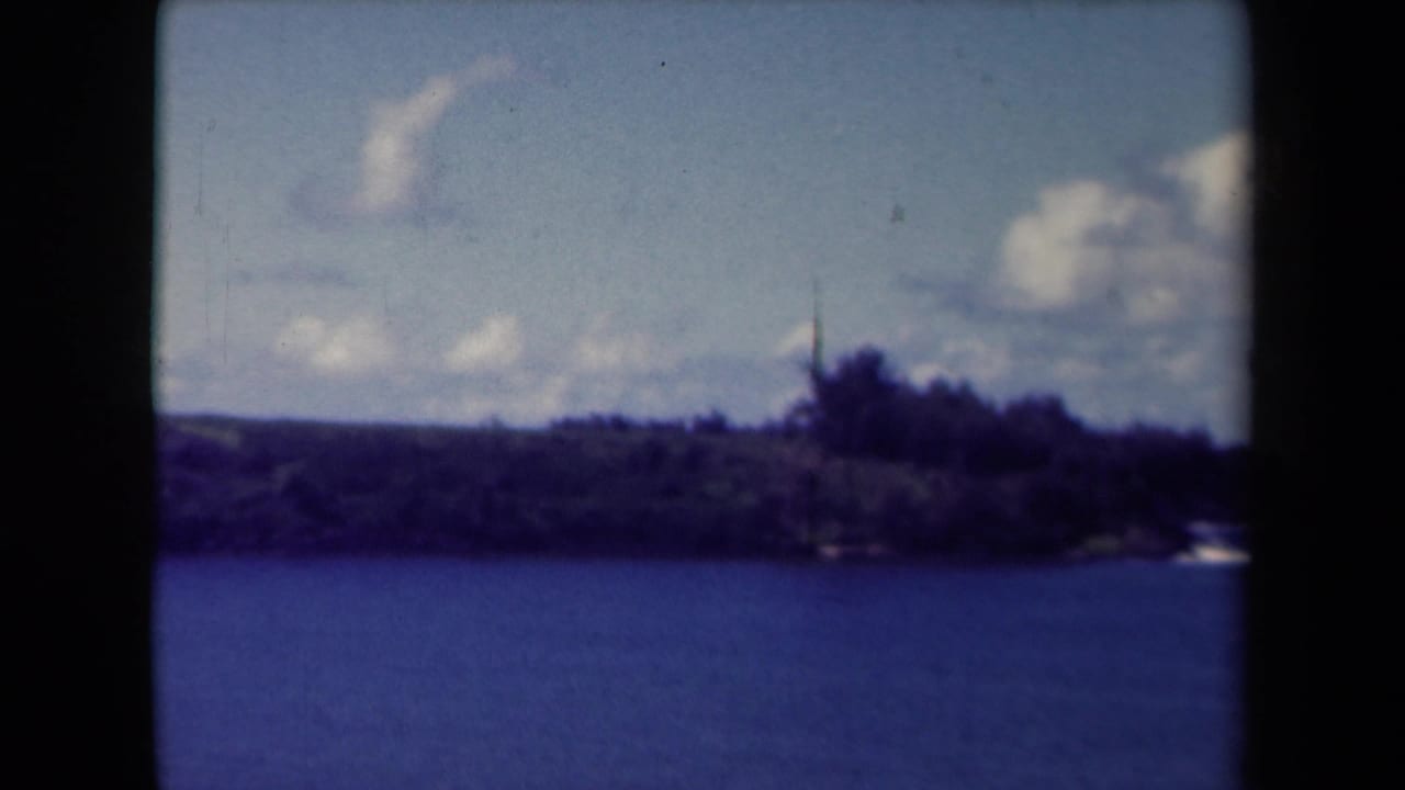 1944 Kauai 8mm view across blue ocean toward a tree-covered shore under scattered clouds.