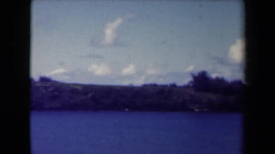 1944 Kauai home-movie frame with ocean foreground, low coastal vegetation, distant hills, and pale clouds.