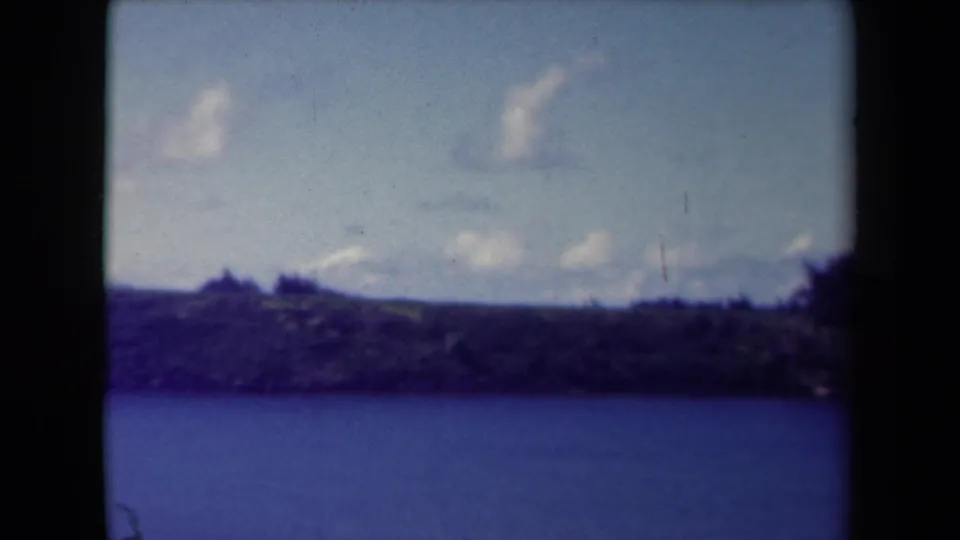 1944 Kauai 8mm frame showing calm ocean, a low wooded shore, distant hills, and scattered clouds near the horizon.