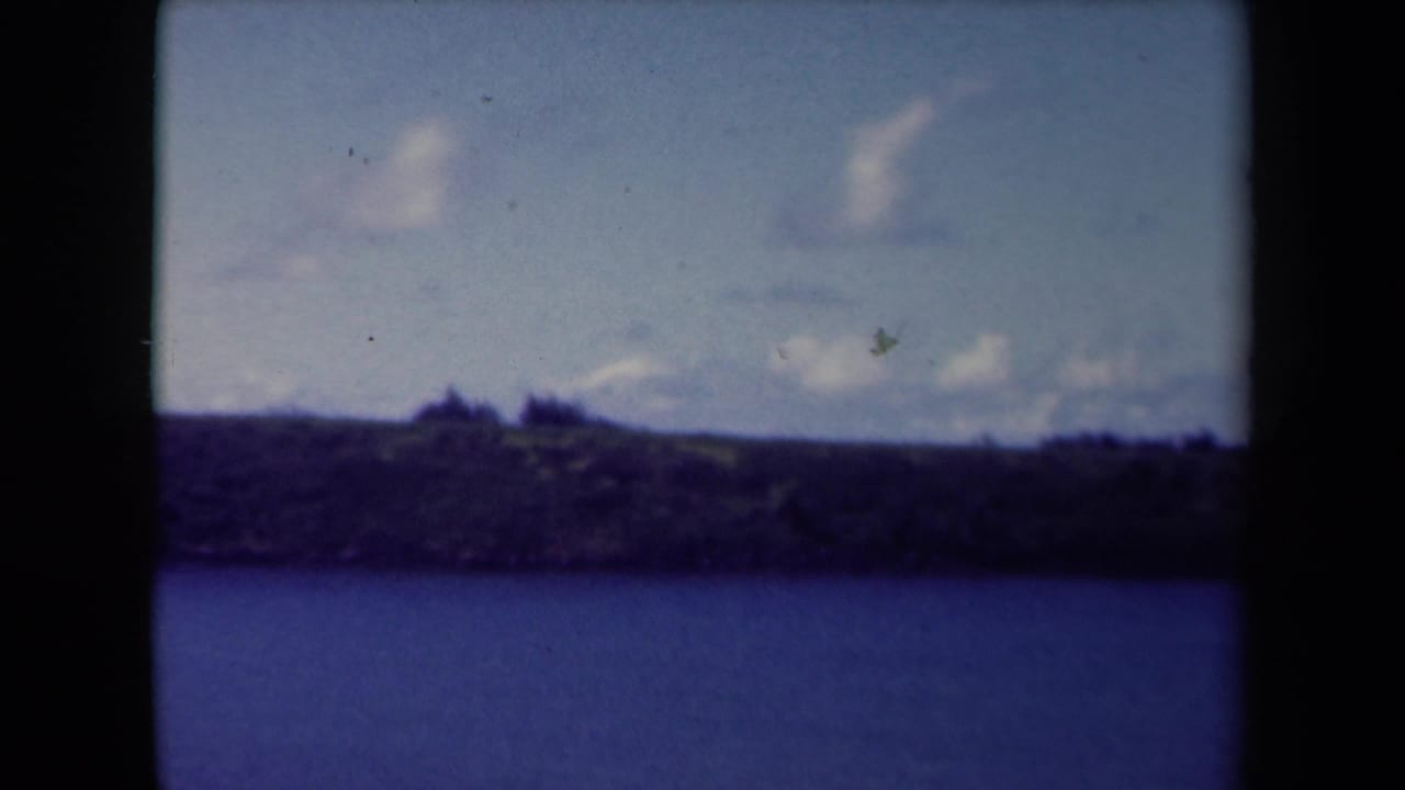 1944 Kauai home-movie frame showing water or shoreline in the foreground with distant low hills and cloudy sky.