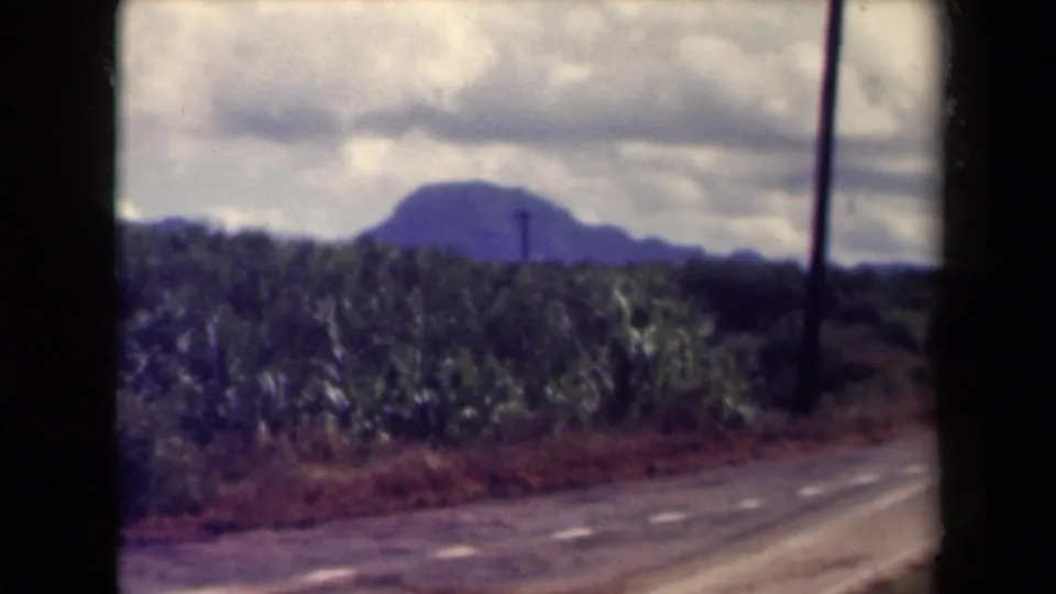 1944 Kauai roadside landscape with green fields, a distant mountain, utility poles, and cloudy sky in soft 8mm color.